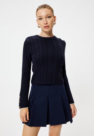 Pull en maille torsadée bleu marine avec un col rond, coupe courte et manches longues ornées de boutons dorés aux poignets, associé à une jupe plissée bleu marine.