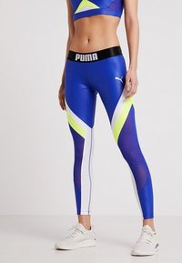 Kvinna som bär blå Puma leggings med vita och neongula accenter, tillsammans med vita träningsskor, stående mot en enfärgad bakgrund.