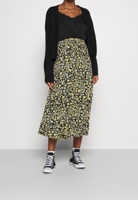 Cardigan a maglia nera, canotta nera e una lunga gonna con un motivo floreale giallo. Abbinato a sneakers alte nere.