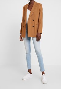 Brun dubbelknäppt kavaj med slag, med svarta knappar och något oversized passform, parat med ljusblå slim fit jeans och vita sneakers.