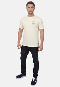 Cremefarvet t-shirt med et motorcykeldesign på venstre bryst, parret med sorte jeans og marineblå sneakers med hvide snørebånd.