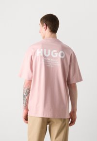 Pink cotton T-shirt with short sleeves, featuring large white "HUGO" text on the back, and text details below. Casual fit and smooth texture.