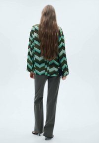 Blusa a righe a zig-zag verde e nera con maniche larghe, abbinata a pantaloni grigi e sandali neri con tacco. Modella di spalle.