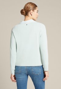 Helles blaues Langarmoberteil mit einem Kragen und einem kleinen Logodetail auf der Rückseite, kombiniert mit blauen Jeans. Weicher Stoff und lässige Passform.