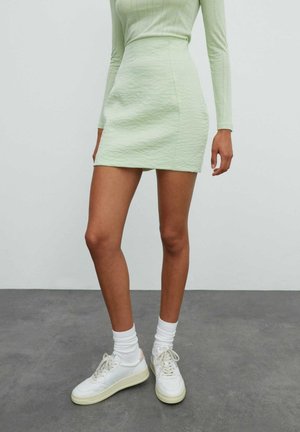 Femme portant une mini-jupe texturée vert clair, un haut à manches longues assorti, des chaussettes blanches et des baskets blanches, debout sur un sol gris.