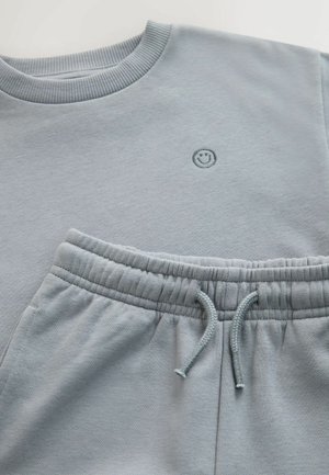 Světle modrá mikina s žebrovaným límcem a malou vyšívanou usmívající se tváří; odpovídající tepláky s elastickým pasem a šňůrkou.