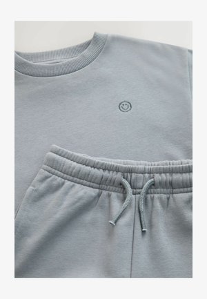 Jasnoniebieska bluza z prążkowanym dekoltem i małą naszywką z uśmiechniętą buźką; do tego pasujące spodnie dresowe z elastycznym pasem i sznurkiem do regulacji.