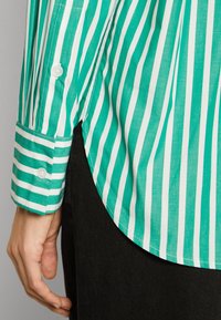 Camicia a righe con linee verticali verdi e bianche, polsino con bottone, orlo curvo e tessuto leggero, indossata sopra un materiale scuro.