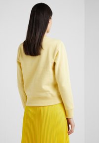 Donna con capelli scuri e lisci, che indossa un maglione giallo pallido e una gonna plissettata gialla brillante, di spalle su uno sfondo bianco semplice.