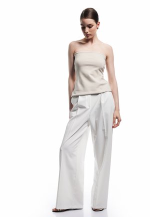 Frau trägt trägerloses beiges Oberteil und weiße, weite Hosen, steht mit gesenktem Kopf vor weißem Hintergrund.