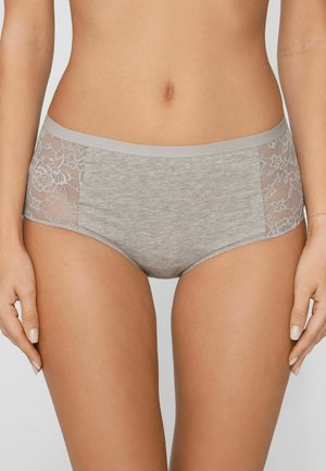 Vrouw die grijze katoenen onderkleding draagt met kanten inzetstukken aan de zijkanten, staand tegen een effen lichte achtergrond.