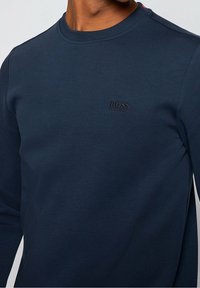 Uomo che indossa una maglietta a maniche lunghe blu navy con girocollo, con un piccolo logo ricamato "BOSS HUGO BOSS" sul lato sinistro del petto.