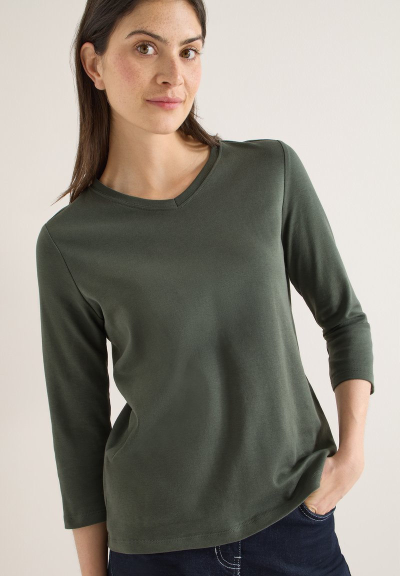 Haut vert foncé à manches longues avec un col en V, fabriqué dans un matériau lisse. Coupe décontractée. Porté avec un jean bleu foncé.