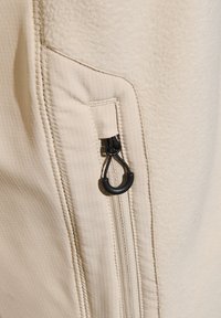 Beige fleecejacka med en texturerad yta, utrustad med en sidazip och en svart ögla för enkel öppning.