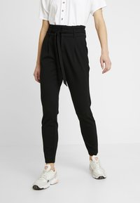 Svarta smala byxor med hög midja och bälte, tillverkade av ett slätt tyg, i kombination med vita och rosa sneakers.