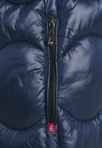 Närbild på marinblå quiltad jacka med dragkedja, rund hundemblem som dragkedjeförsedd och en liten röd hundbricka nedanför dragkedjan.