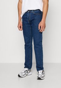 Mørkeblå denimjeans med slim fit, med fem lommer og søm-detaljer, kombinert med hvit T-skjorte og grå sneakers.