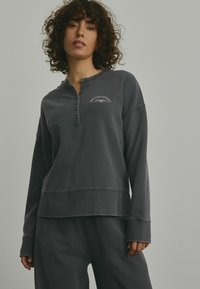 Mørkegrå pullover sweatshirt med knaplukning ved halsen, ribbede manchetter og et dekorativt tryk med teksten "NEW YORK CITY" på brystet. Blød tekstur.