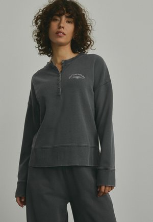 Mørkegrå pullover sweatshirt med knaplukning ved halsen, ribbede manchetter og et dekorativt tryk med teksten "NEW YORK CITY" på brystet. Blød tekstur.