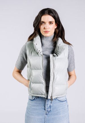 Lichtgrijze puffer vest met hoge kraag, ritssluiting en drukknoopjes. Gequilte ontwerp, zijzakken, gedragen over een grijze trui.