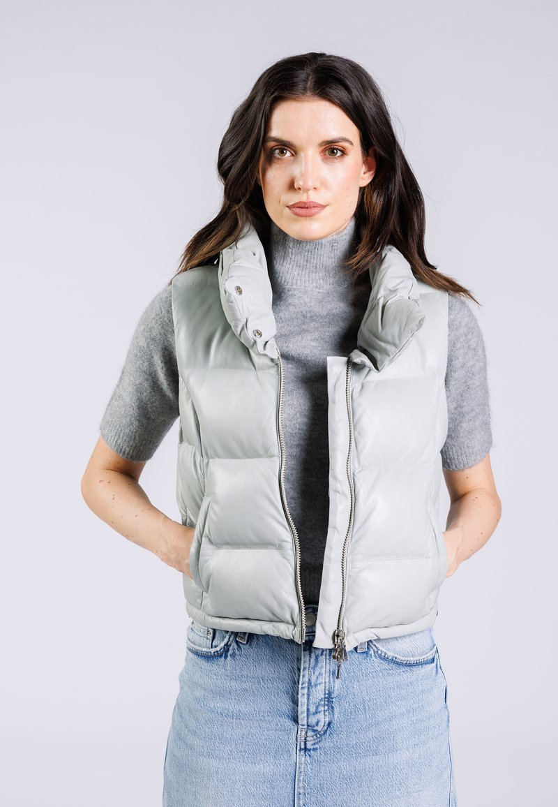Gilet trapuntato grigio chiaro con collo alto, chiusura a zip e bottoni a pressione. Design trapuntato, con tasche laterali, indossato sopra un maglione grigio.