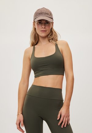 Kvinde iført olivengrøn sports-bh og leggings samt en taupefarvet baseballkasket, står i et neutralt studie og kigger mod kameraet.