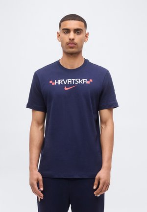 Mann mit kurzen Haaren, der ein dunkelblaues T-Shirt mit "HRVATSKA" und einem roten Nike-Swoosh über der Brust trägt, steht vor einem einfarbigen Hintergrund.