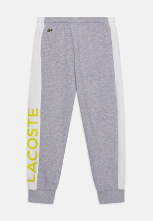 Pantalon de survêtement gris avec un panneau latéral blanc arborant le mot "LACOSTE" en jaune vif. Fabriqué en tissu doux, avec une taille élastique.
