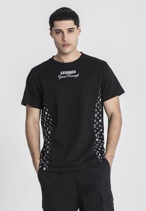 Jeune homme aux cheveux courts et foncés portant un t-shirt noir Kavanagh avec des panneaux latéraux blancs à motifs et un pantalon cargo noir, les mains dans les poches.