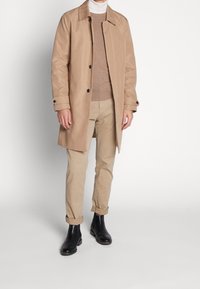 Trench-coat beige avec un col à revers, détails à boutons, pull léger, pantalon kaki et bottines noires, debout sur un fond neutre.