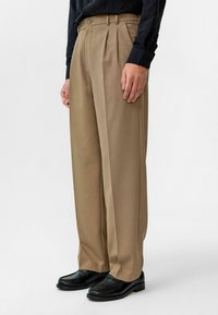 Beige geplooide broek met een getailleerde pasvorm, gemaakt van soepel stof. Gecombineerd met zwarte instappers, wat een klassieke, verfijnde uitstraling geeft.