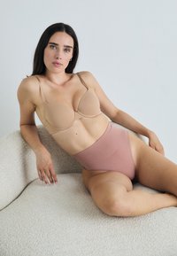 Beige polstret bh med justerbare stropper og høyt liv rosa undertøy, begge laget av glatt stoff. Modell ligger på en teksturert hvit sofa.