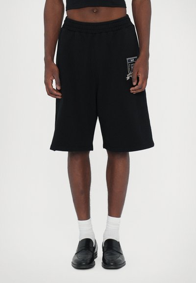 Sorte bomulds shorts med elastisk talje, med en sidelomme med lap. Båret med sorte slip-on sko og hvide sokker.