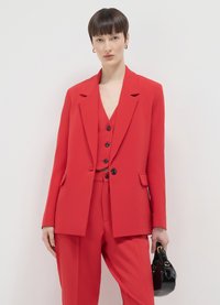 Blazer rosso sartoriale con gilet abbinato, caratterizzato da bottoni neri e tasche. Abbinato a pantaloni rossi e una piccola borsa nera.