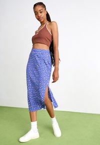 Fustă midi florală albastră cu slituri laterale, combinată cu un top scurt maro și adidași albi, pe un fundal verde.
