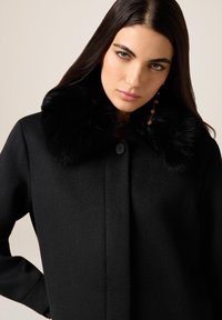 Cappotto in lana nera con colletto in morbido ecopelliccia nero, chiusura con un solo bottone e texture liscia. Design semplice e strutturato.