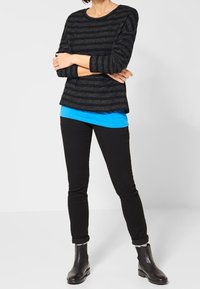 Svartrandig tröja med metalltrådar över en blå topp, kombinerad med svarta smala jeans och svarta ankelskor.