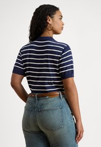 Sieviete ar cirtainiem matiem, valkā zelta stīpu auskarus, tumši zilu un baltu svītrainu īsroku topu, brūnu jostu un gaiši mazgātas džinsas, stāv sāniski.