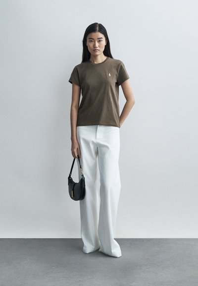 Barna pamut rövid ujjú póló kerek nyakkivágással, széles fehér nadrággal párosítva. A modell egy kis fekete kézitáskát tart a kezében.