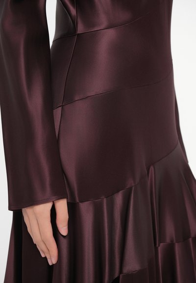 Burgundska satenska obleka z dolgimi rokavi, z večplastnimi volani na spodnjem robu in gladko teksturo. Na zgornjem delu so vidni šivi.