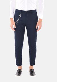 Chinos blu navy con una texture liscia, taglio tapered, orli arrotolati e un dettaglio di catena argentata sulla tasca. Indossati con scarpe slip-on nere.