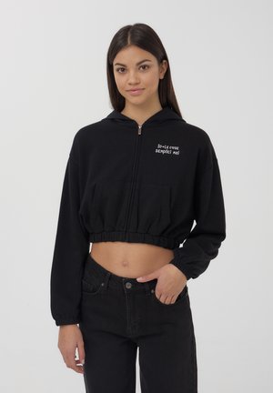 Felpa nera corta con zip anteriore, maniche lunghe, orlo elastico e tasca frontale con testo bianco. Abbinata a jeans neri.
