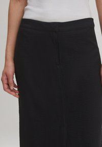 Jupe noire texturée, longueur genou, avec une taille lisse et des détails de couture sur le côté, portée avec un haut blanc partiellement visible.