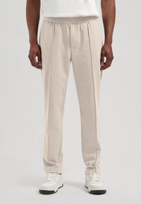 Lyst beige bukser laget av mykt stoff, med elastisk midje og fremre folder. Kombinert med hvite joggesko for kontrast.