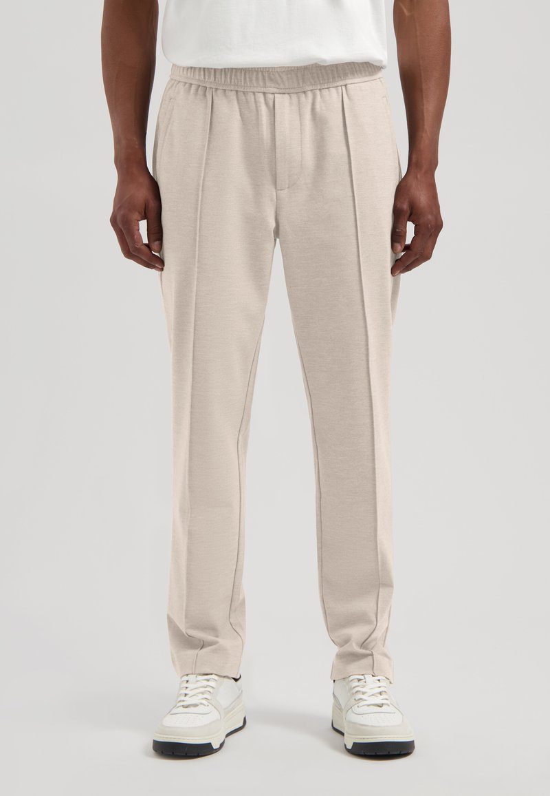 Lyst beige bukser laget av mykt stoff, med elastisk midje og fremre folder. Kombinert med hvite joggesko for kontrast.