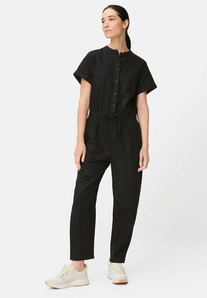 Kvinde står iført en sort jumpsuit med knapper, korte ærmer, talje, der er indsnævret, og hvide sneakers foran en ensartet lys baggrund.