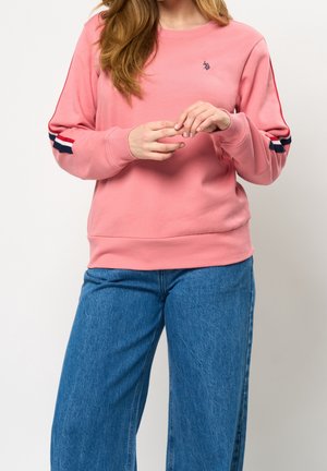 Vrouw in een roze sweatshirt met rode, witte en marineblauwe strepen op de mouwen en een blauwe spijkerbroek, handen voorzichtig voor zich aanraakend.