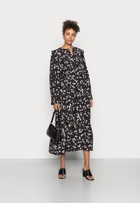 Vestido maxi preto com estampado floral, mangas longas, design em camadas, gola e pequenos padrões florais em branco e rosa. Combinado com saltos bloco pretos.