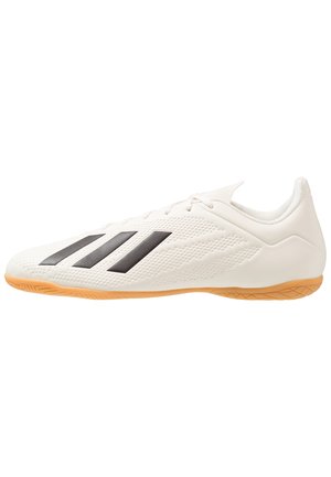 Chaussure de football en salle blanche avec des rayures noires sur le côté et une semelle en gomme marron clair.