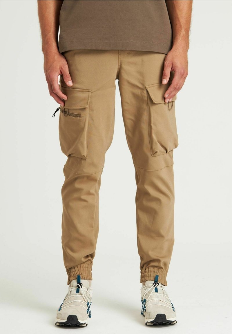 Homme portant un pantalon cargo beige avec plusieurs poches et des baskets blanches avec des touches de bleu, debout devant un fond uni.